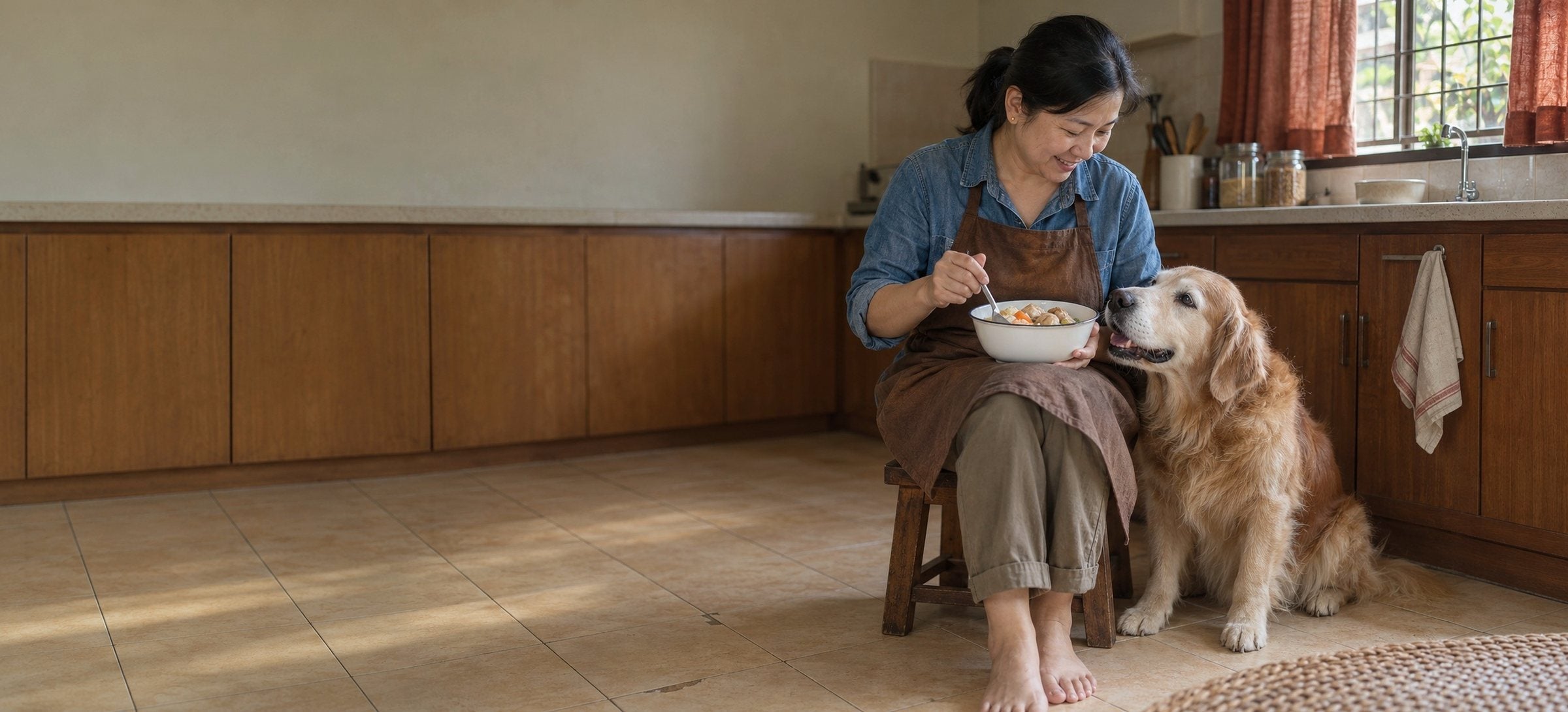 A woman on a low kitchen stool stirring food in a bowl, her old golden retriever sitting close beside her and looking up at her